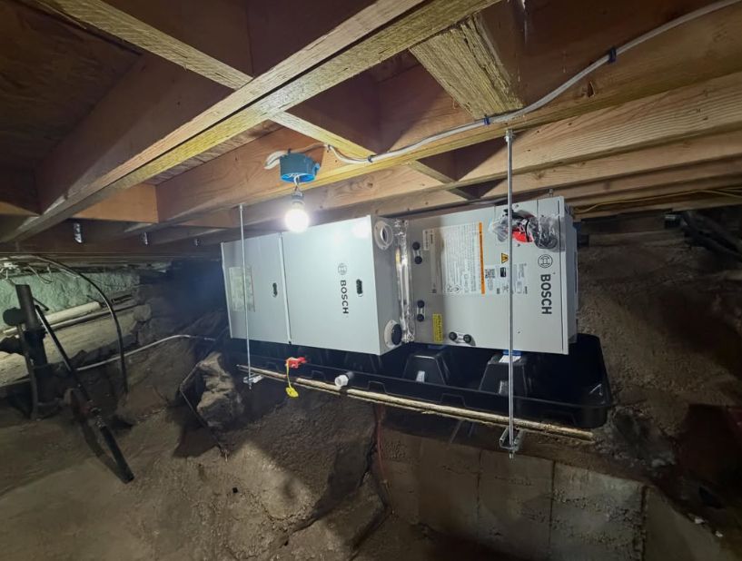 Furnace and HVAC unit installed in a crawlspace under exposed wooden beams, lit by a single bulb.