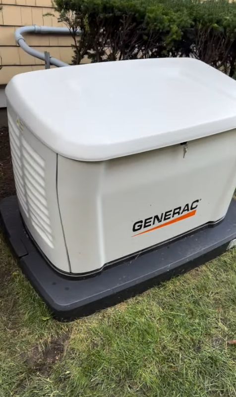 White Generac generator on a black base beside a house, with a hose and shrubbery in the background