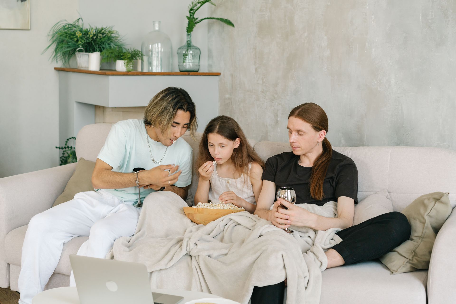 Three people on a couch looking at a laptop, with blankets, mugs, and houseplants in a cozy living room.