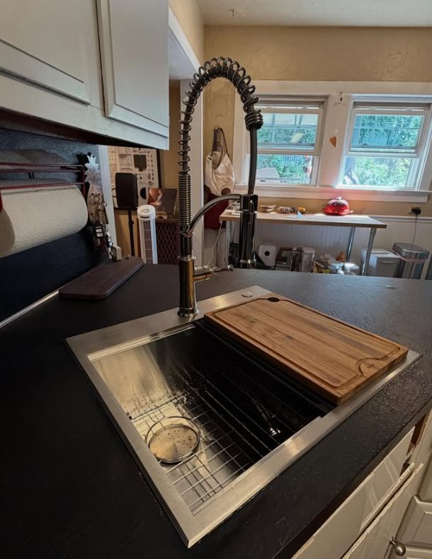 Kitchen sink with high-arc faucet and wooden cutting board, beside a window.