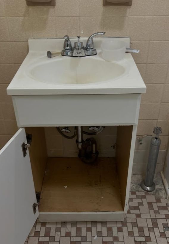 White bathroom vanity with sink, open cabinet door, exposed plumbing, and patterned tile floor.