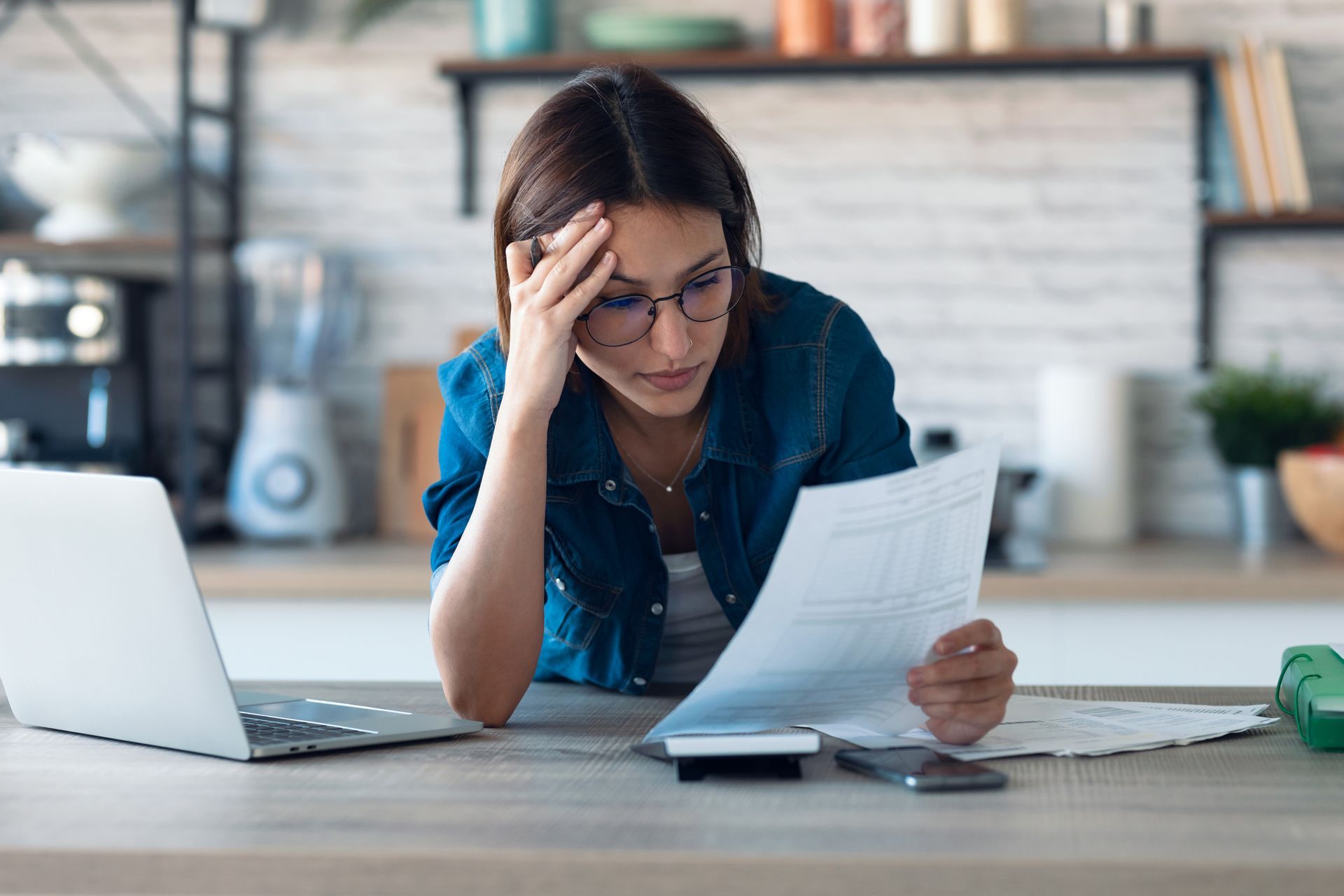 Small business owner reviewing bookkeeping and financial paperwork at a laptop, looking stressed.