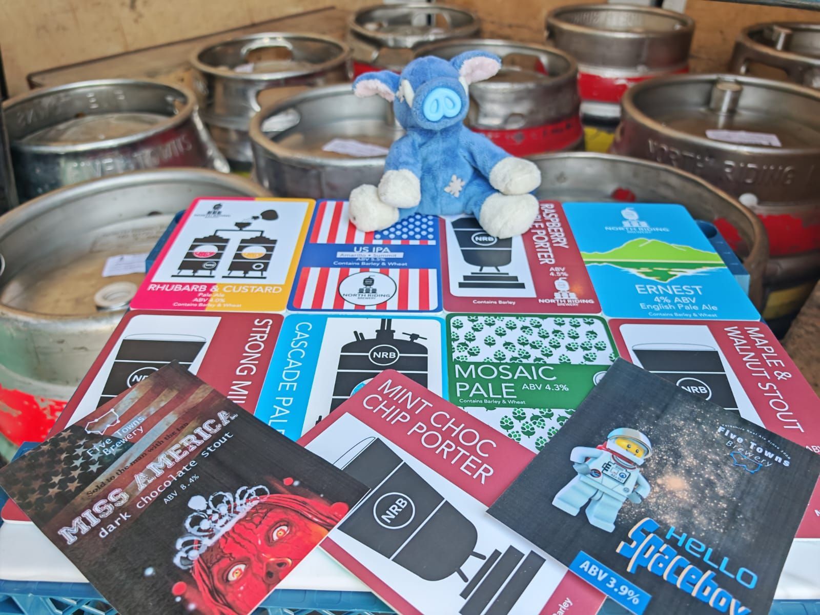 Plush donkey on beer labels atop crates, surrounded by kegs. Labels include various beer names and graphics.