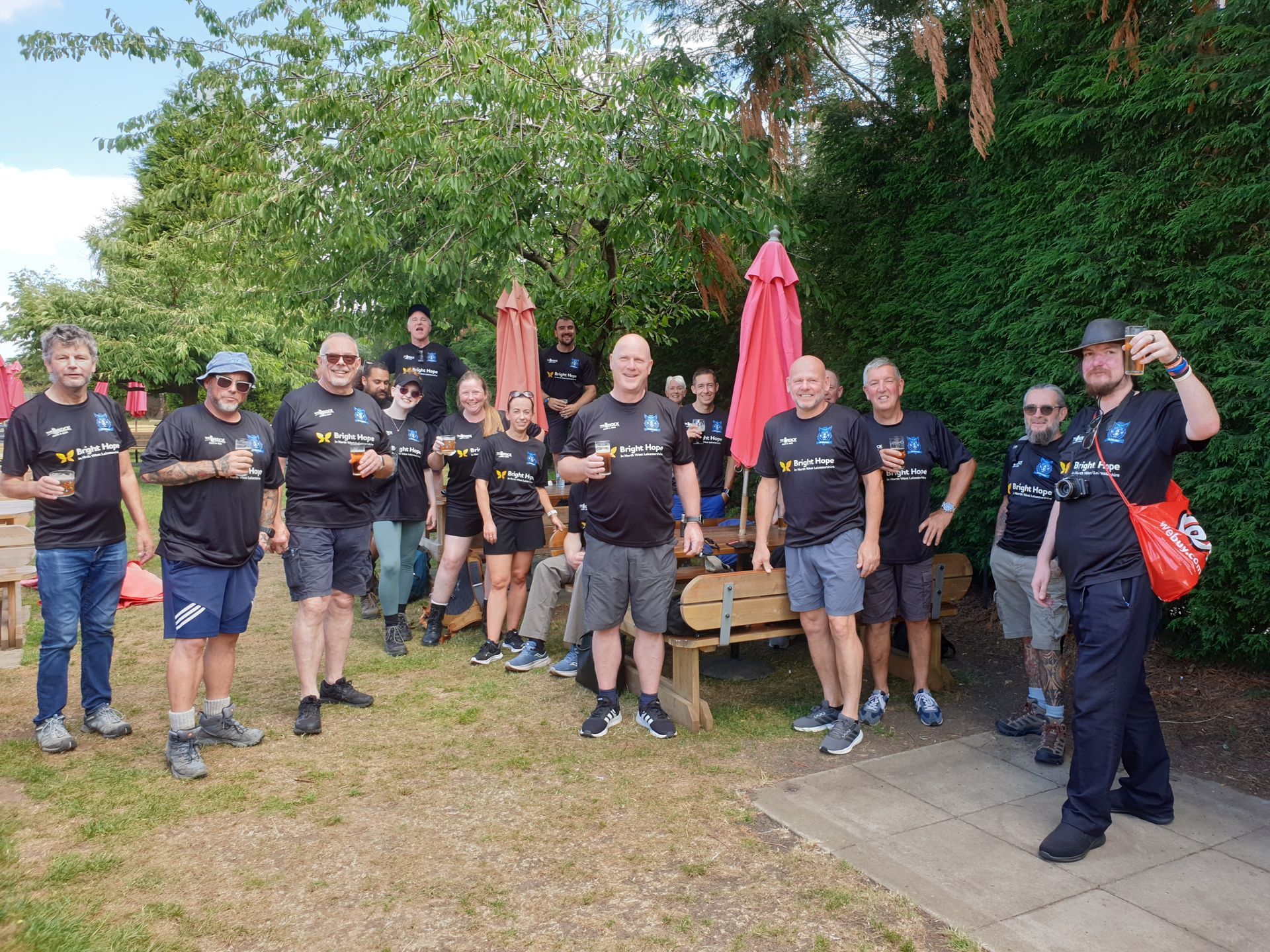 Group of people in black shirts pose outdoors near a picnic table, holding drinks and smiling.