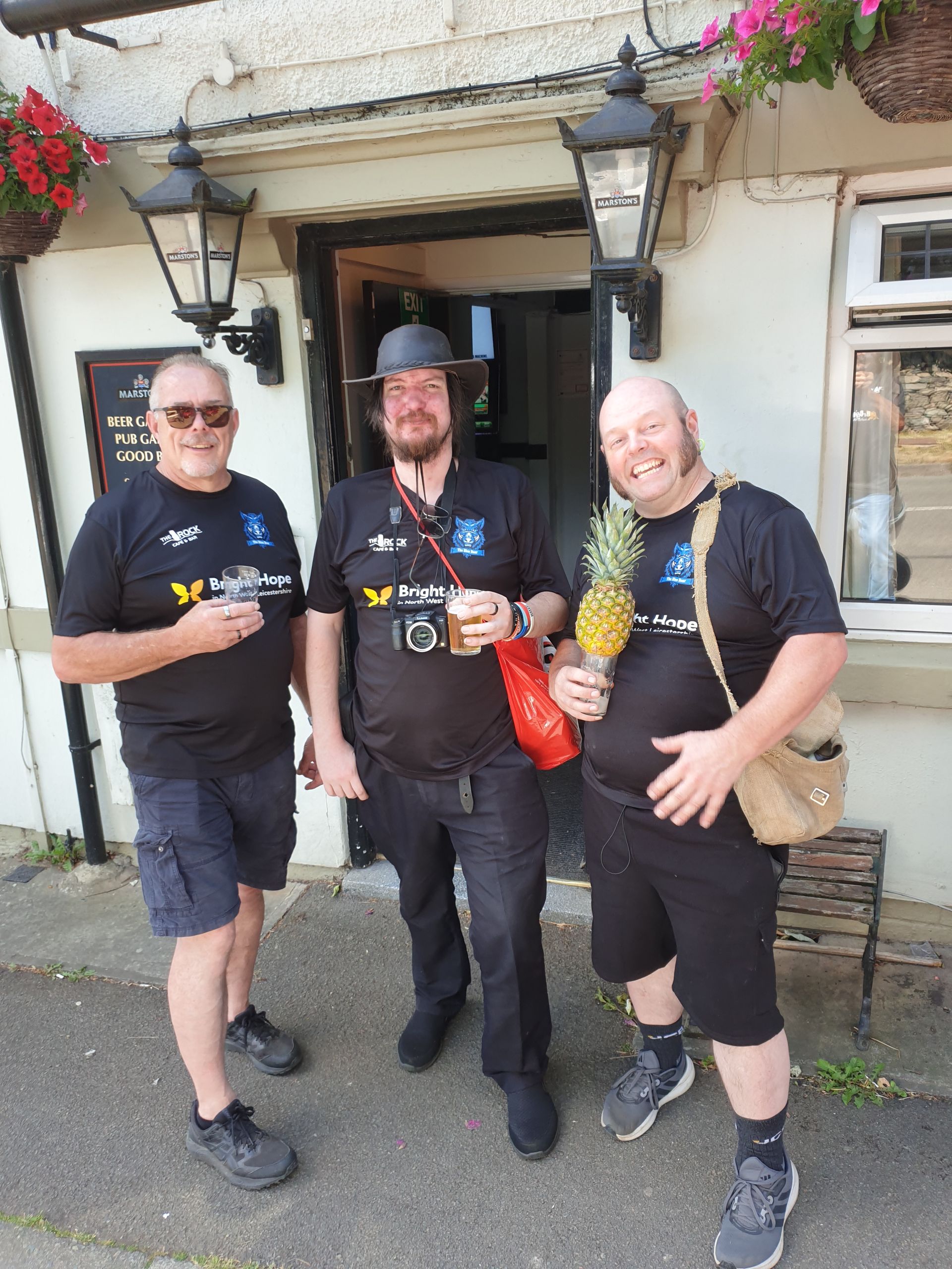 Three men in black t-shirts outside a pub. One holds a pineapple, all are smiling.