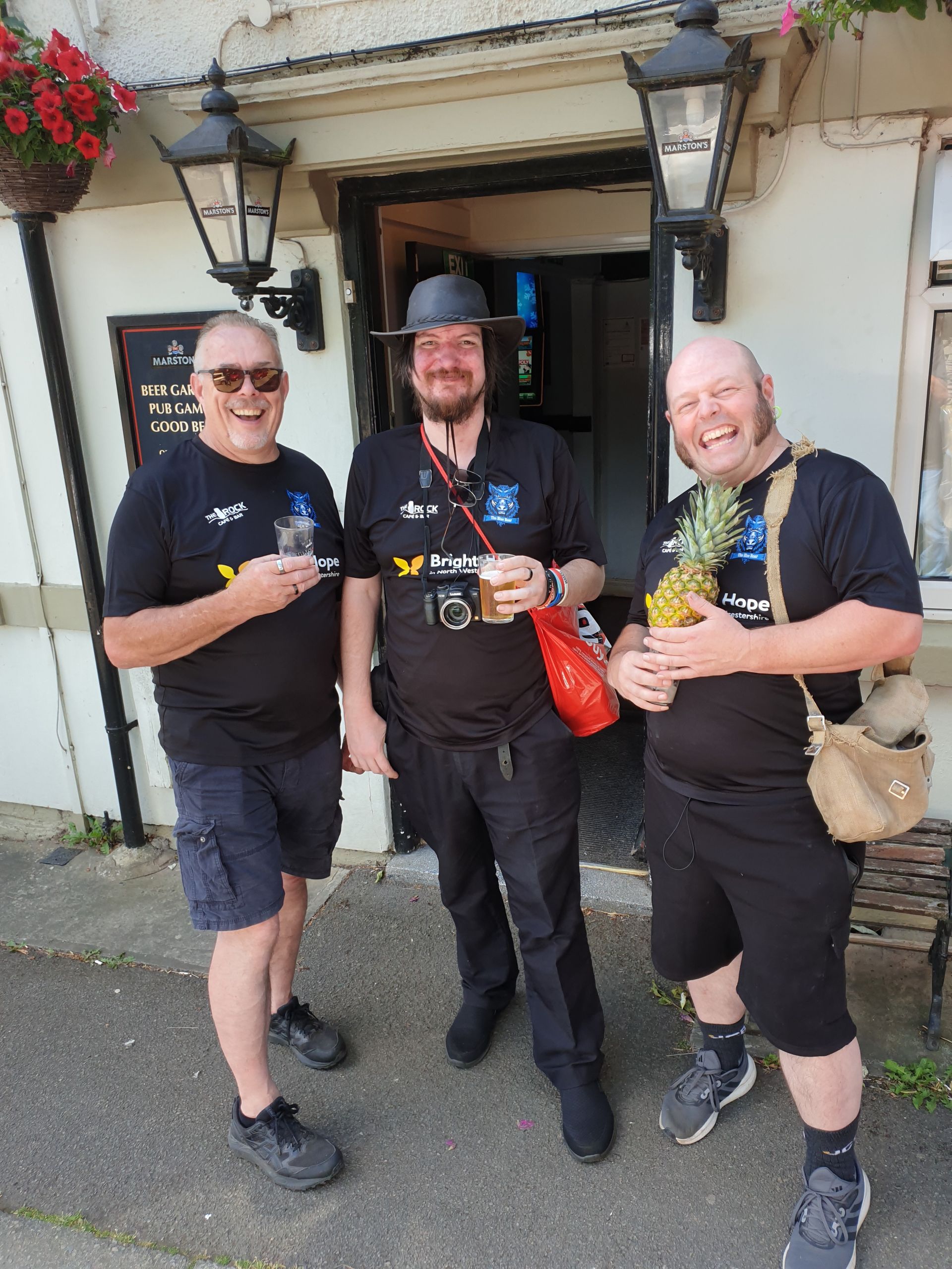 Three men outside a building, wearing black shirts. One holds a pineapple, another a drink. They smile.