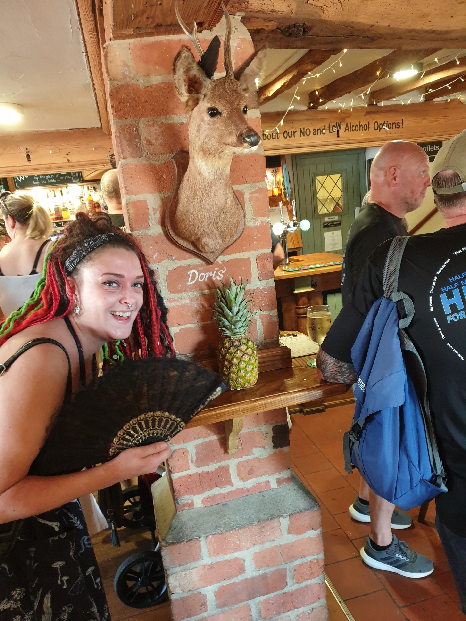 Woman with red-streaked hair holds fan, smiles near a brick column with a pineapple and taxidermied deer head.
