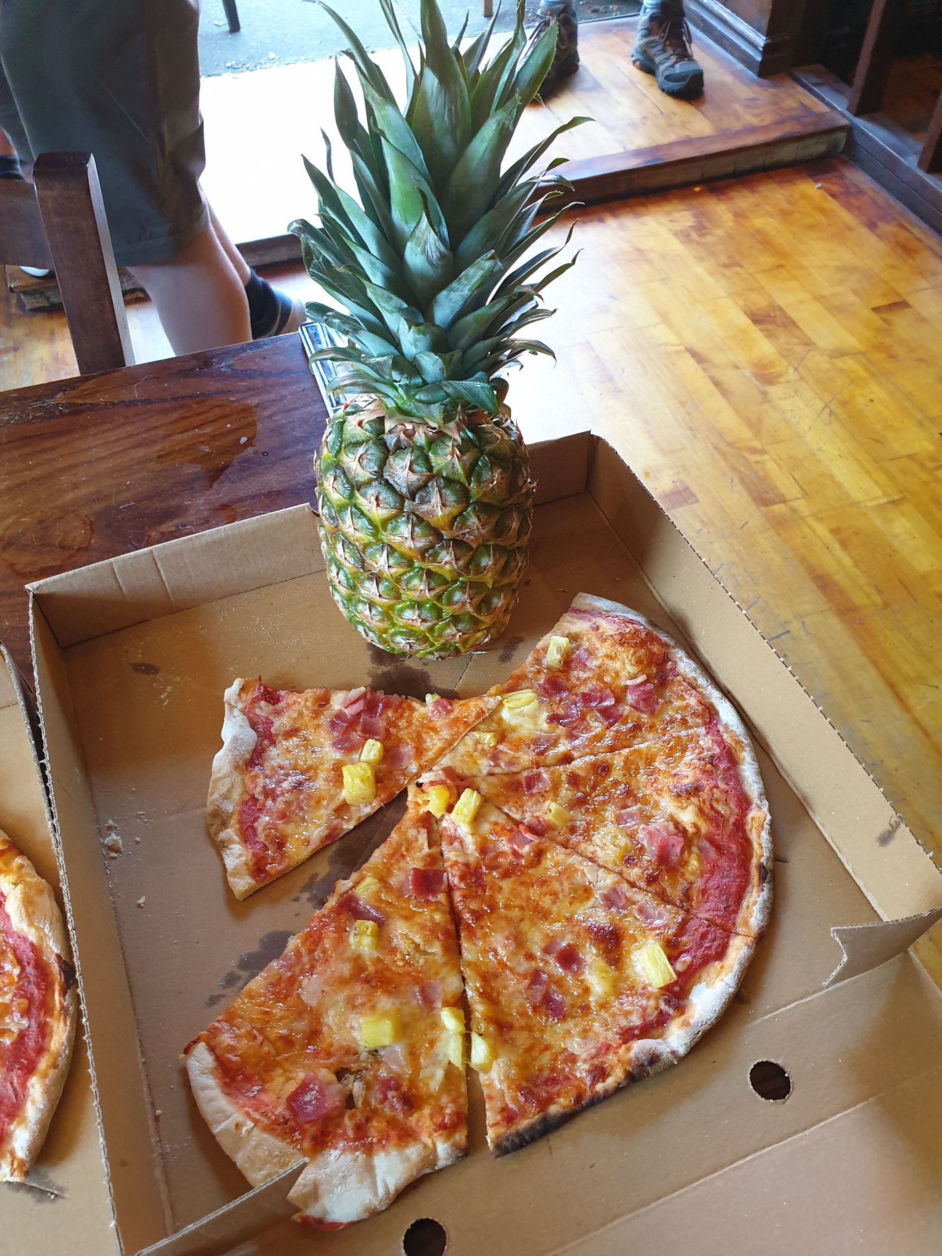 Pineapple and a pizza with pineapple slices in a cardboard box, on a wooden surface.