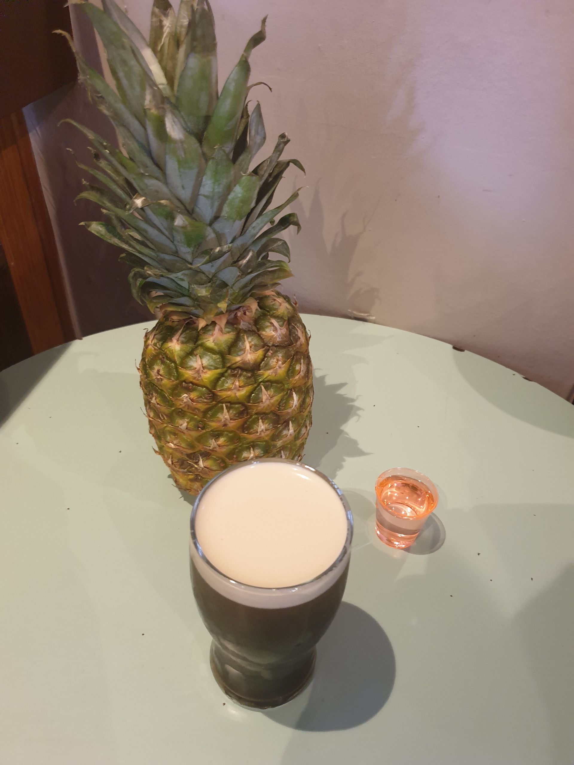 Pineapple beside a glass of dark liquid, and a small orange cup on a pale green surface.
