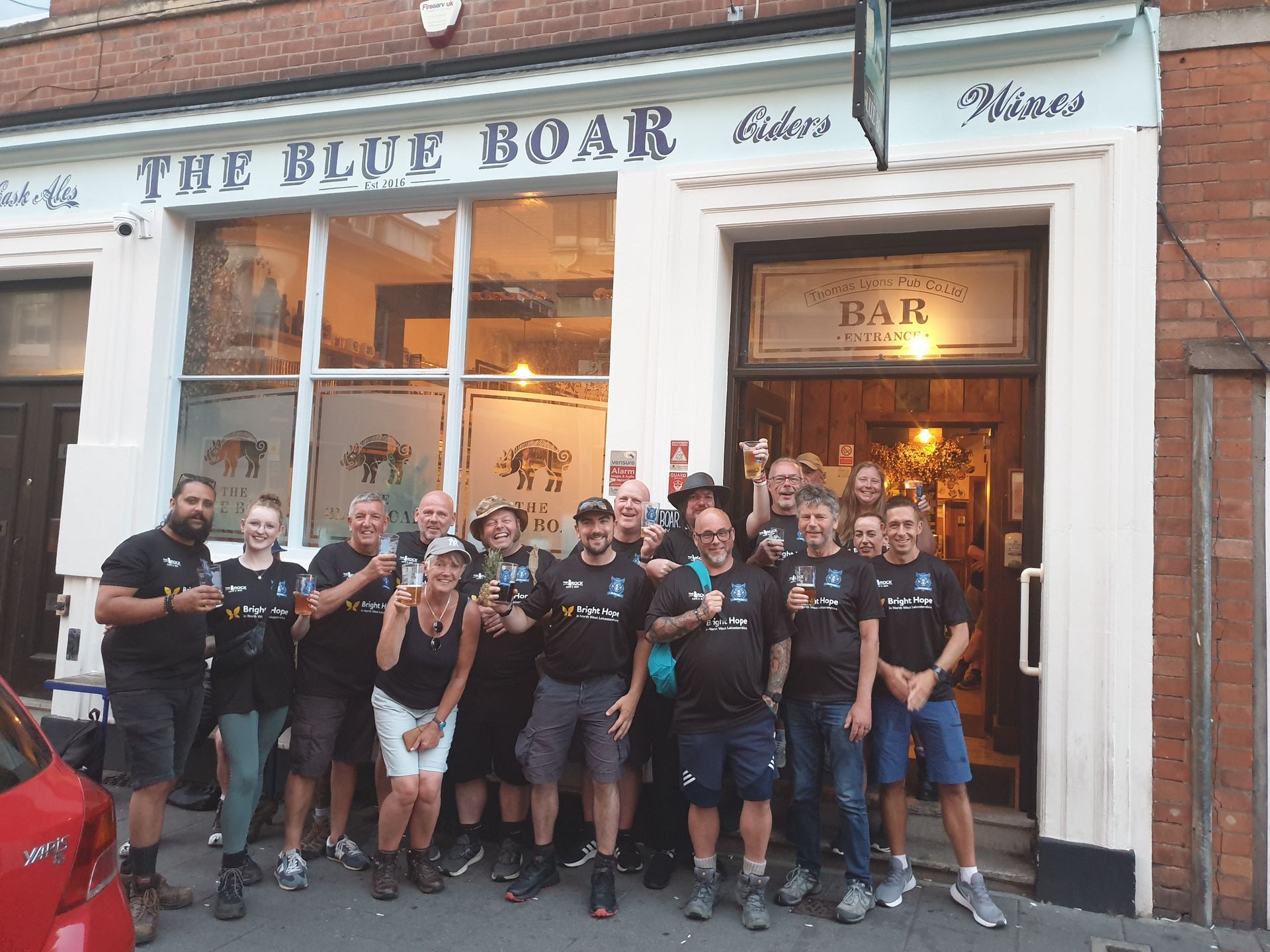 Group of people toasting outside the Blue Boar pub with drinks, smiling, in front of the entrance.