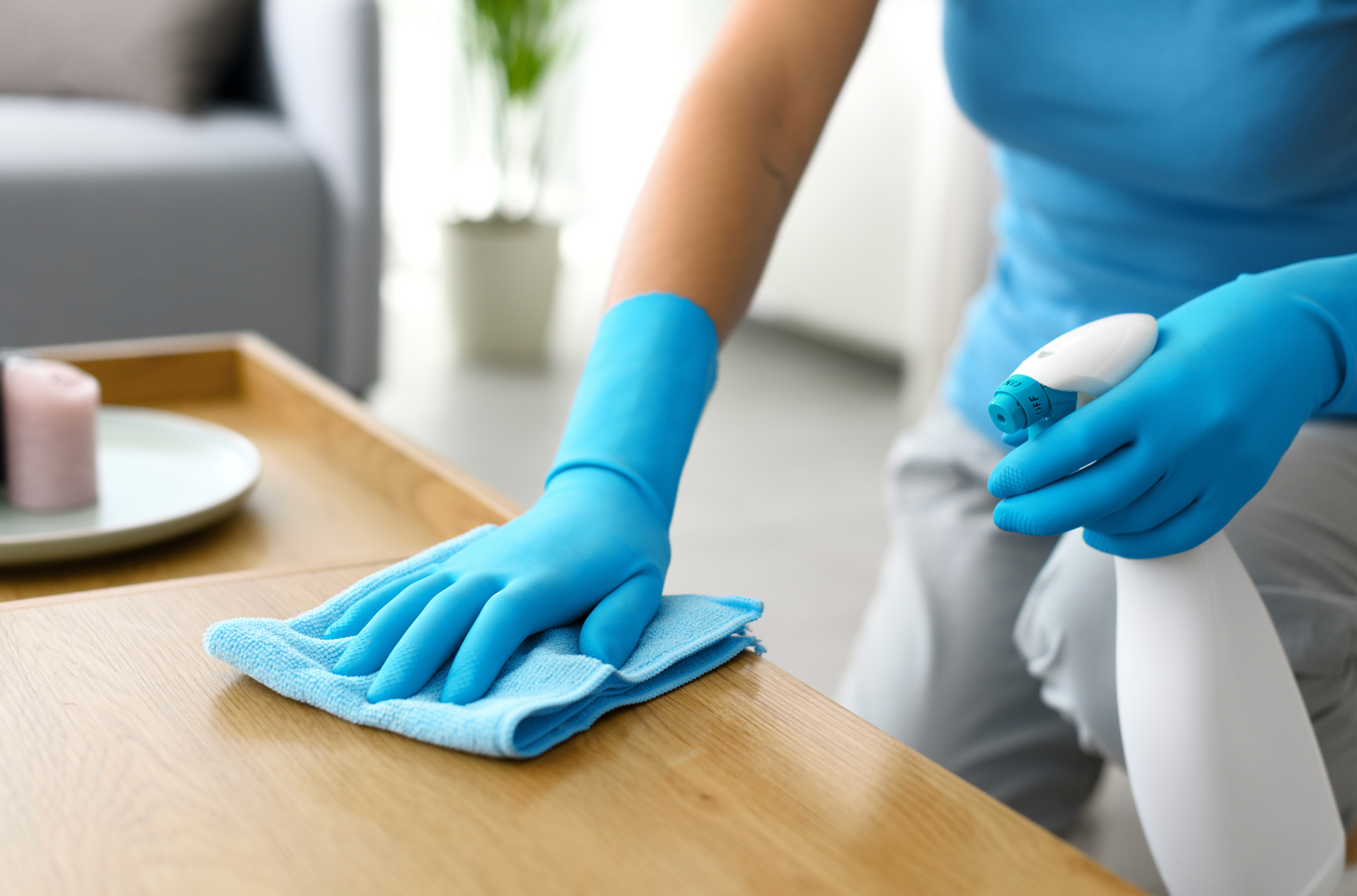 A woman in blue gloves is cleaning a wooden table with a cloth and spray bottle.