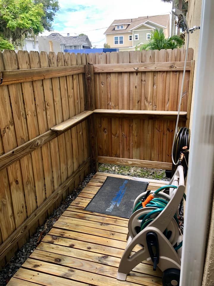 Enclosed wooden outdoor shower with a wooden floor and fence. A hose reel, mat, and shelf are visible.