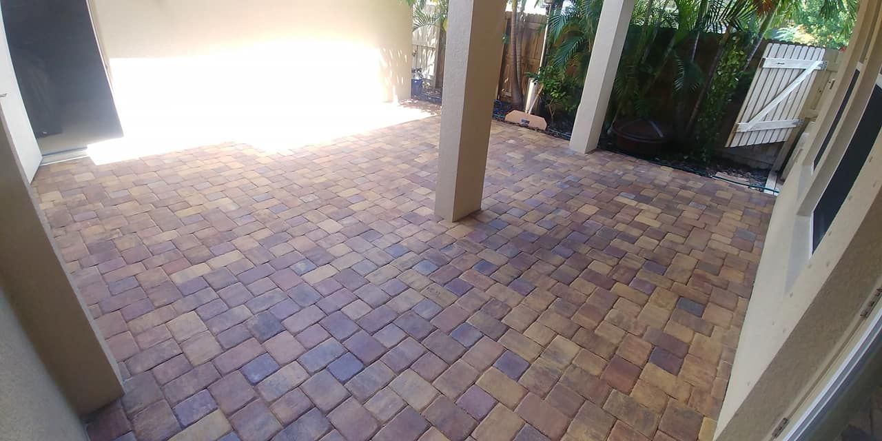 Patio paved with multi-colored bricks, surrounded by columns and foliage. Bright sunlight illuminates the open space.