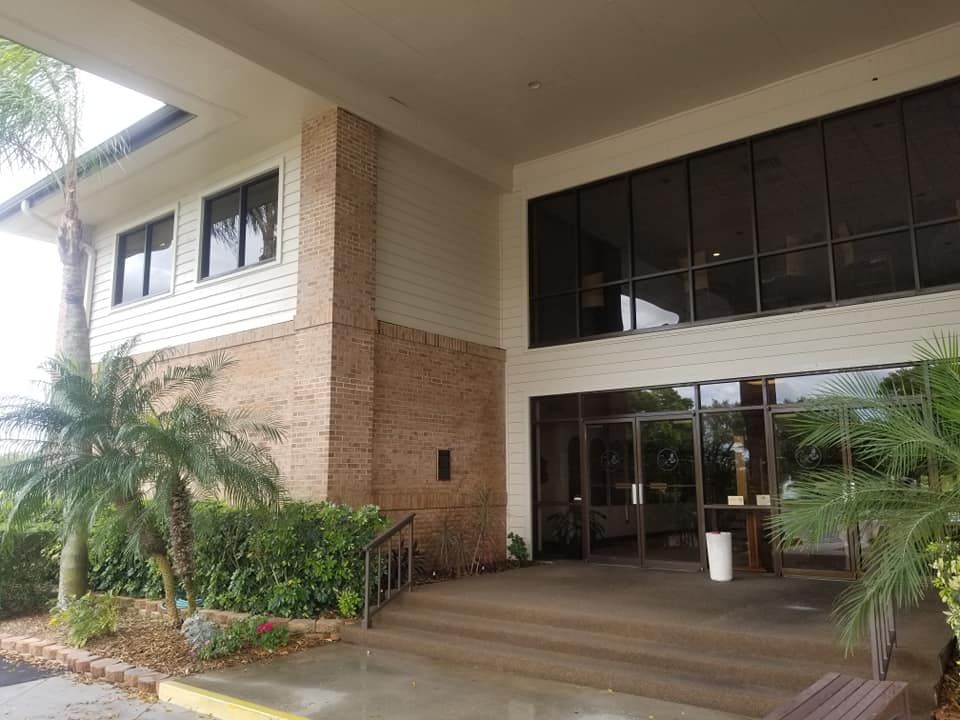 Entrance to a building with brick and glass. Steps lead to the doorway, with palm trees and greenery in the foreground.