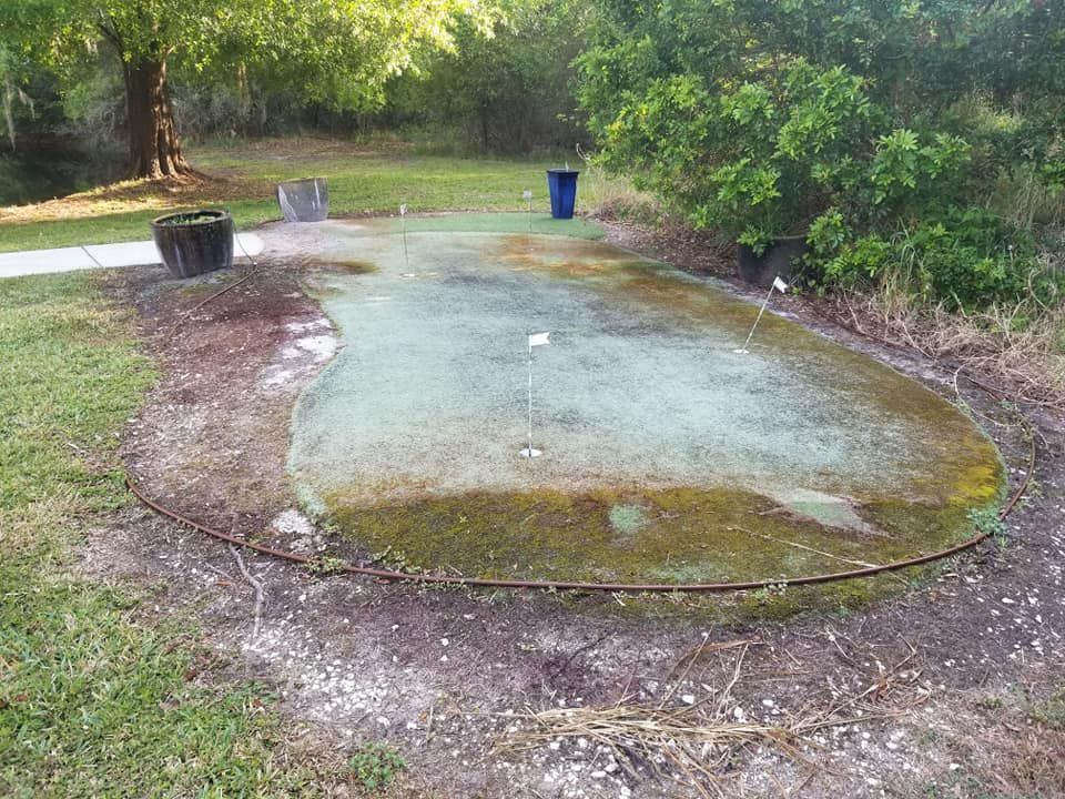 Dilapidated putting green with moss, pots, and a trash can in a park setting.