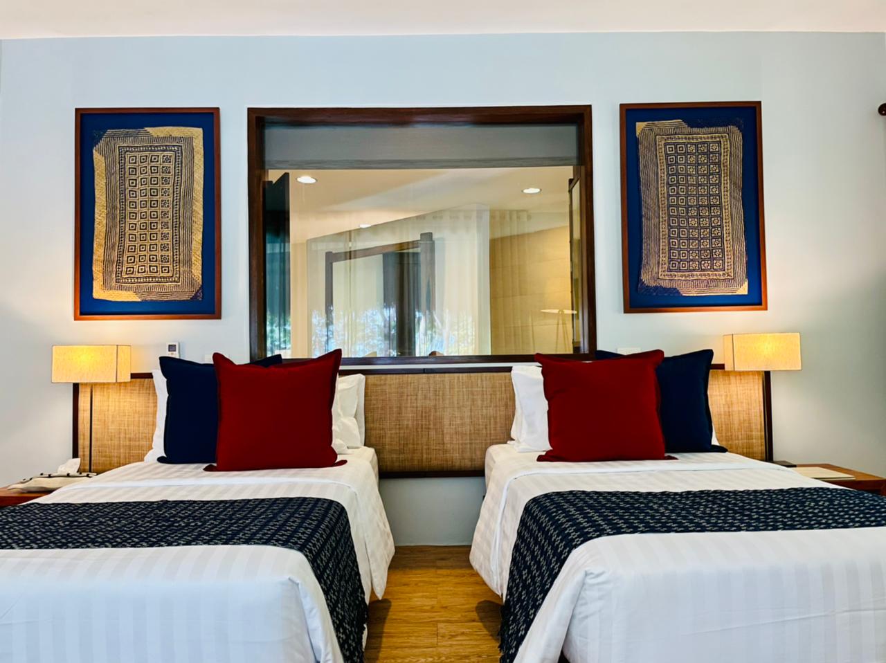 Twin beds in a modern hotel room with blue accents, red pillows, lamps, and framed wall art.