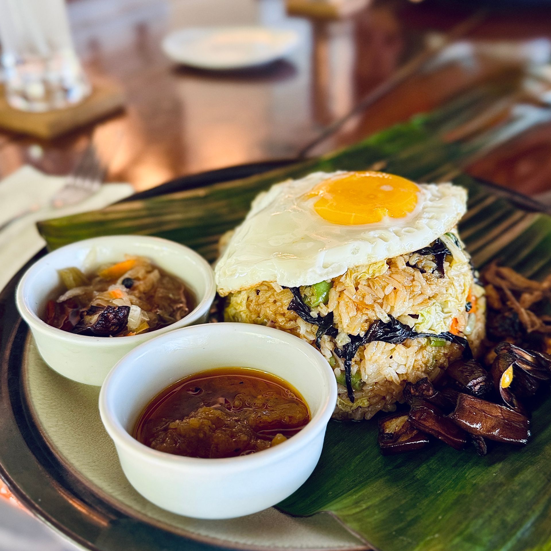 Plate of fried rice topped with a fried egg, served on a banana leaf with two dipping sauces