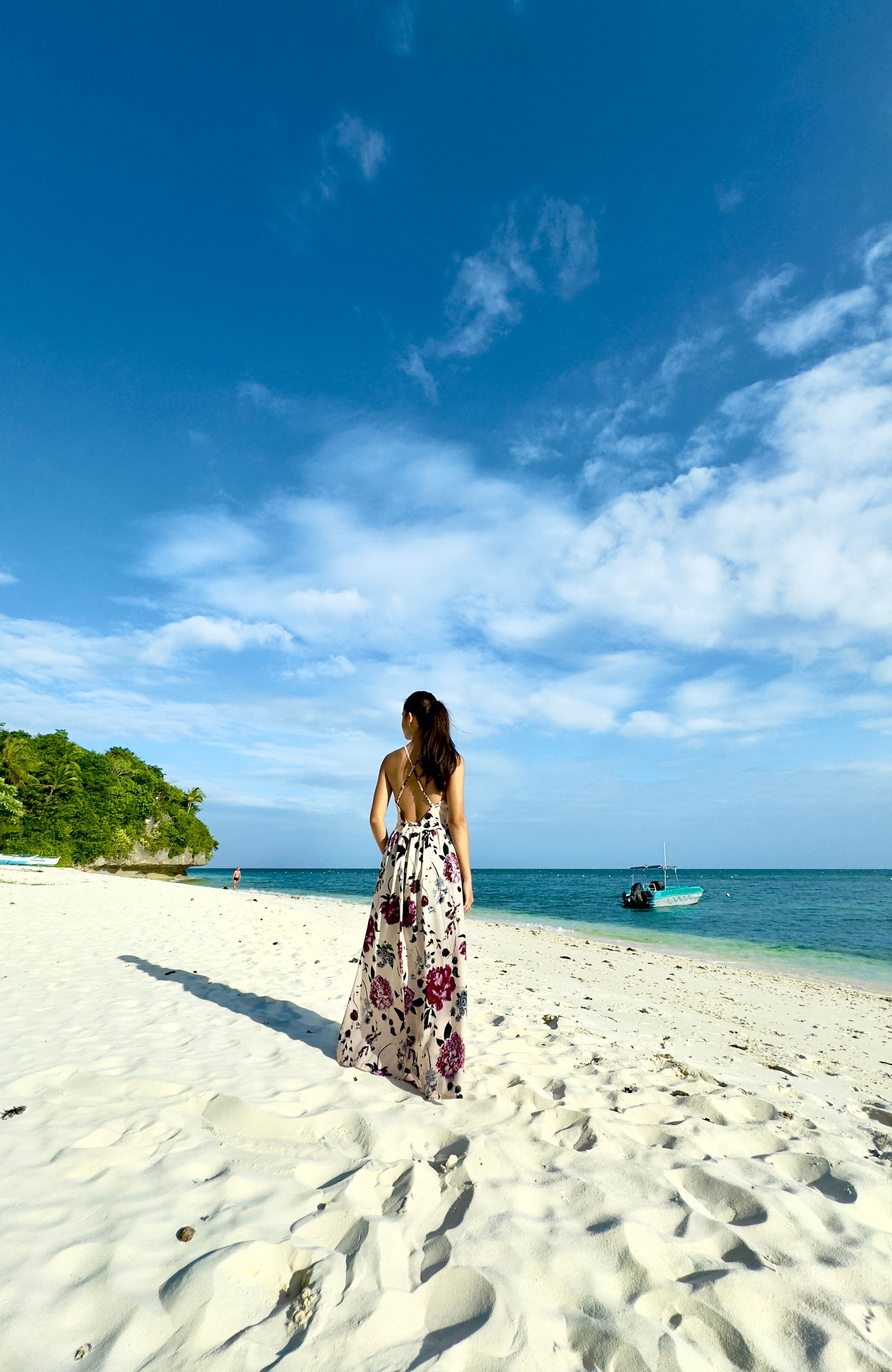Person in a floral dress walking on a sandy beach under a bright blue sky.