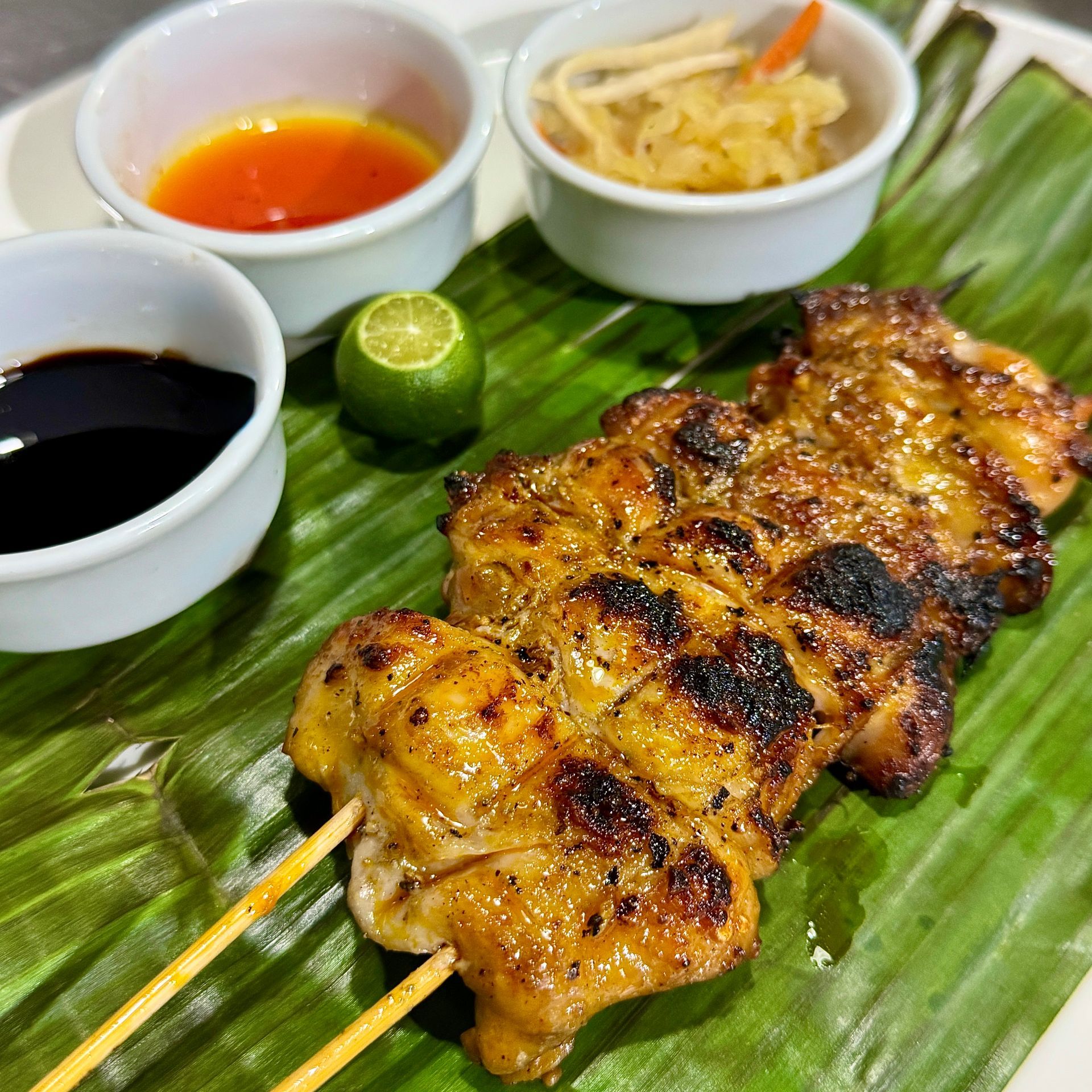 Grilled chicken skewers on banana leaf with dipping sauces, lime, and pickled vegetables