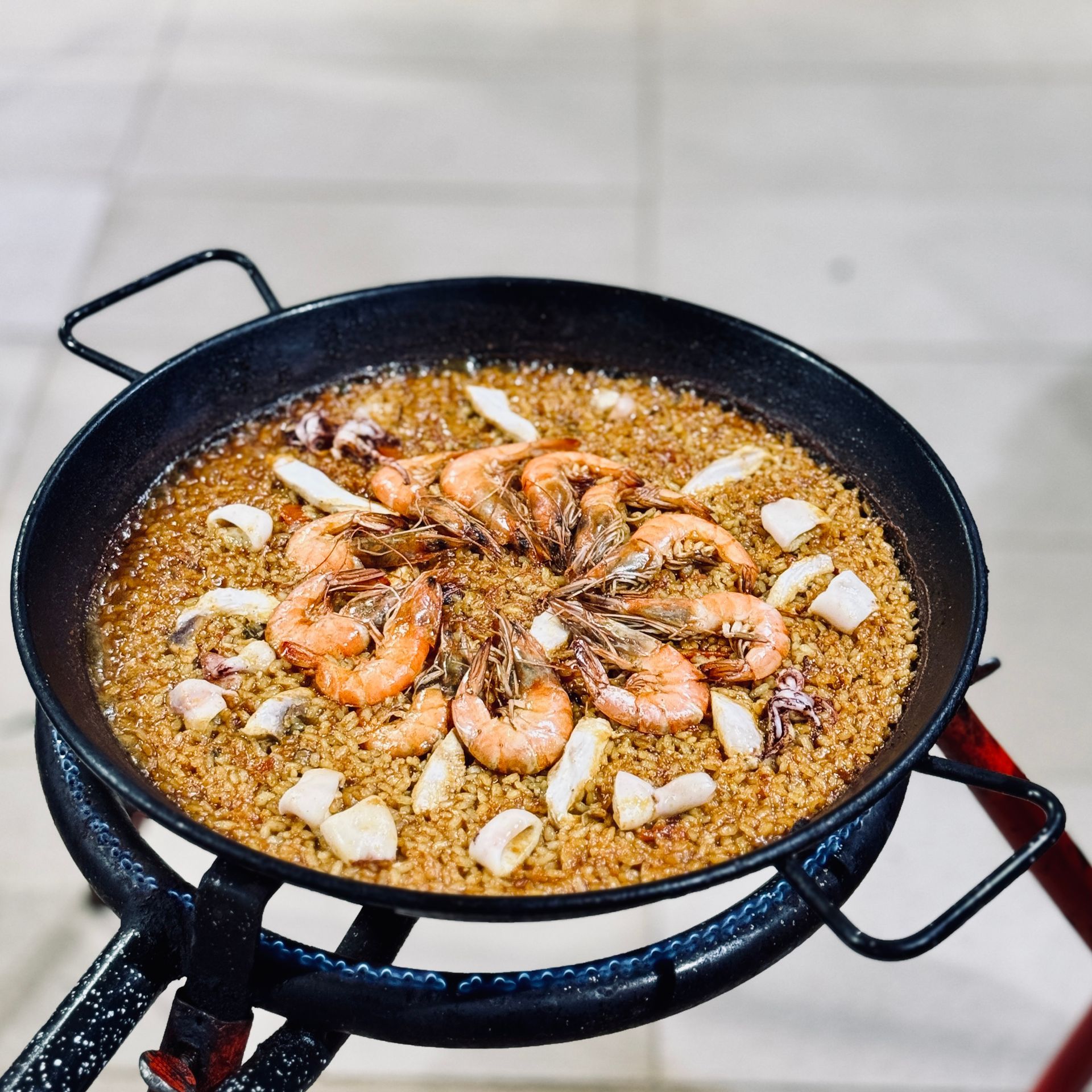 Paella pan with shrimp and shellfish on a stovetop, topped with sliced onions and herbs