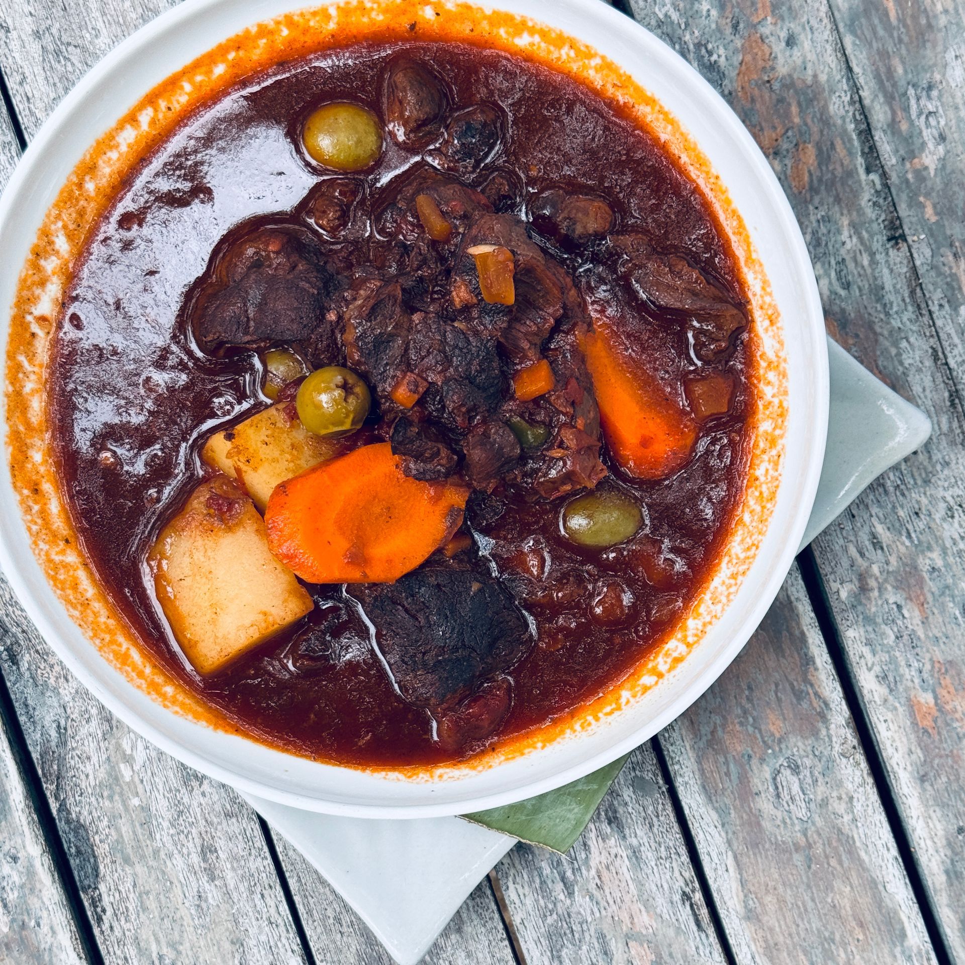 Bowl of dark beef stew with carrots, potatoes, and olives on a wooden table