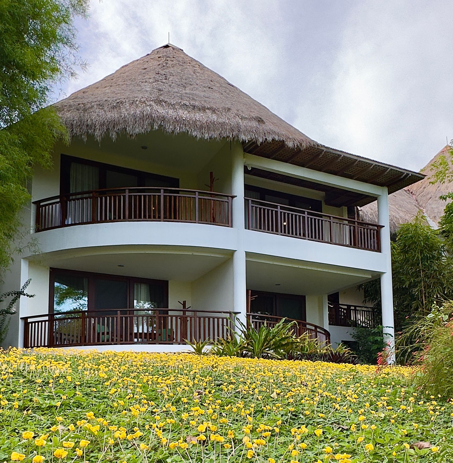 Two-story tropical villa with thatched roof, wooden balconies, and yellow flowers in front