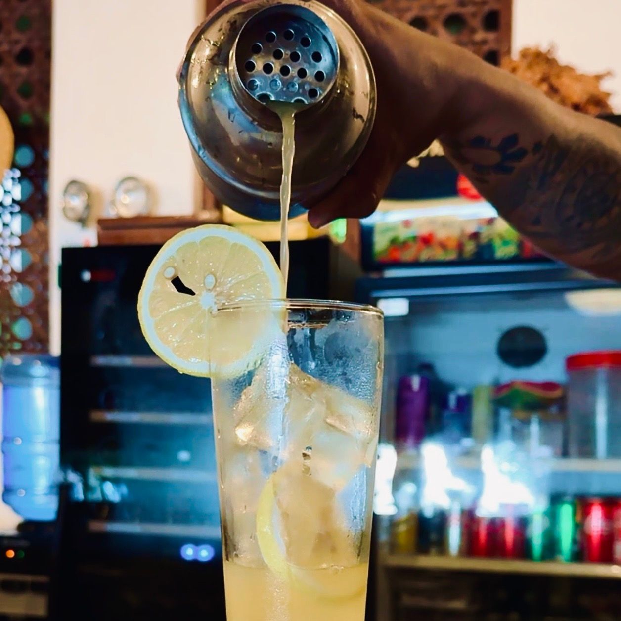 A hand pours a drink into a tall glass with ice and a lemon slice.