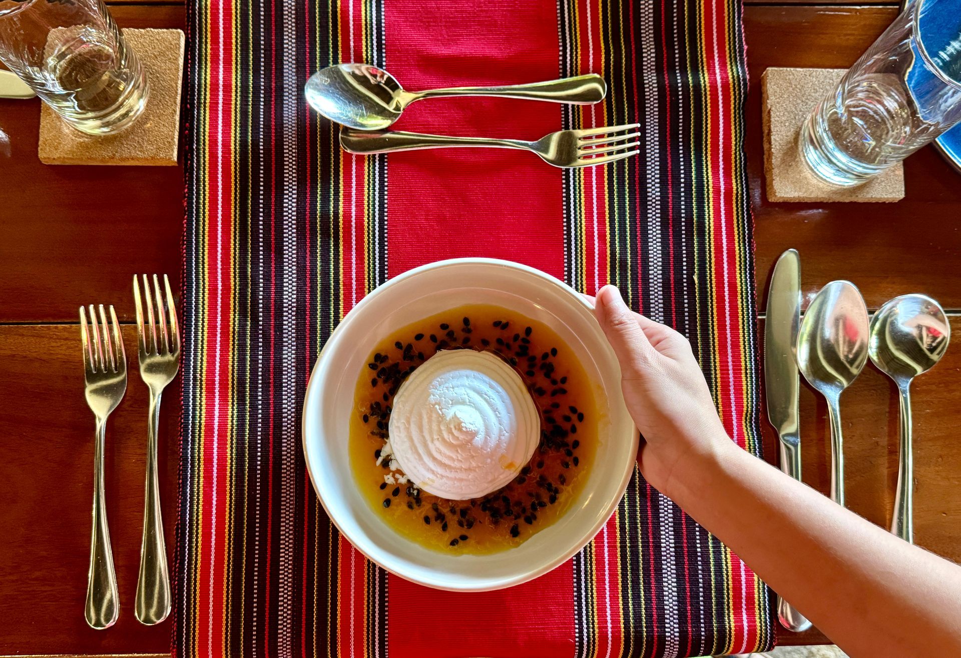 Overhead view of a breakfast table with a bowl of yogurt, red striped tablecloth, and a child’s hand reaching in