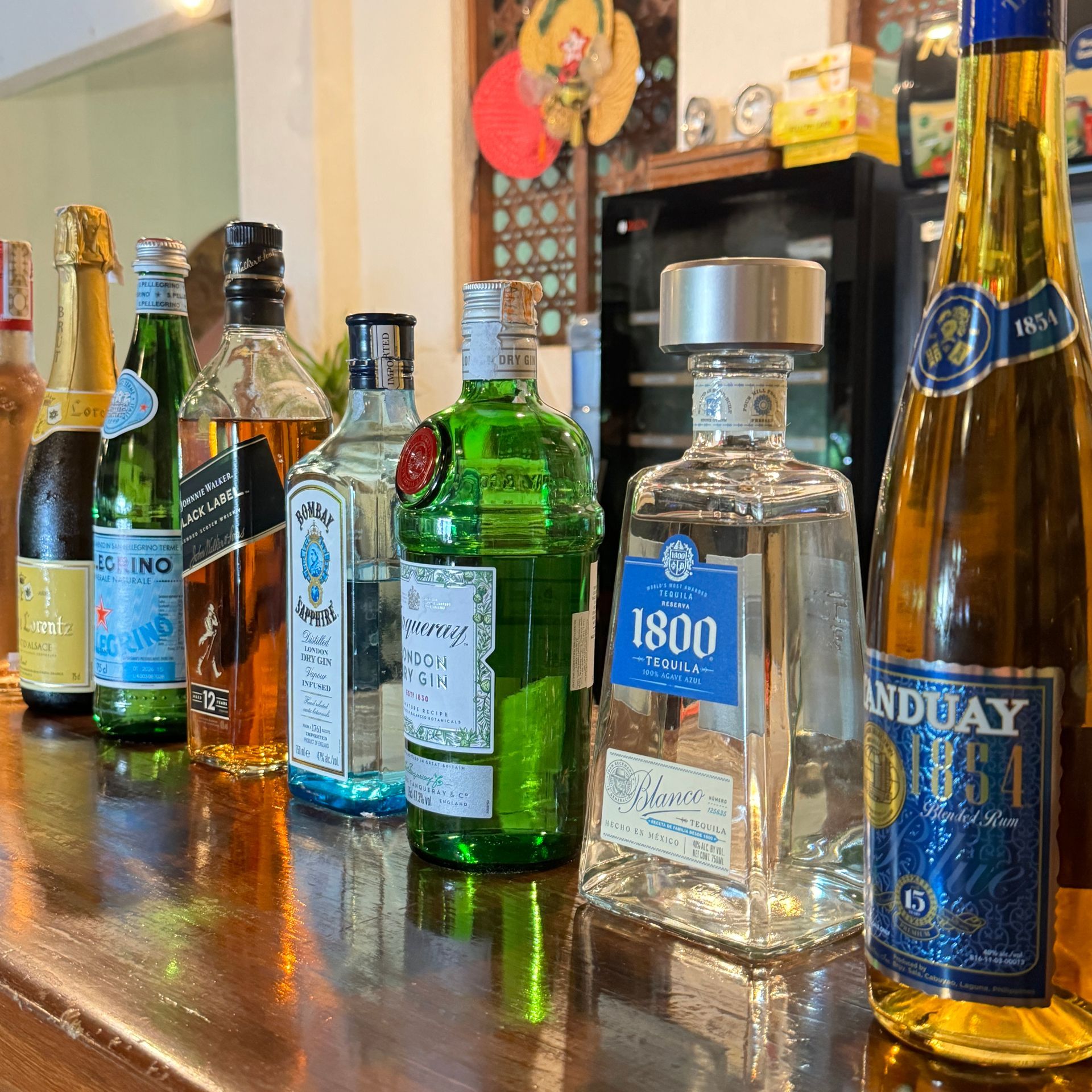 Assorted liquor bottles lined up on a wooden bar counter, including green, clear, and blue bottles.