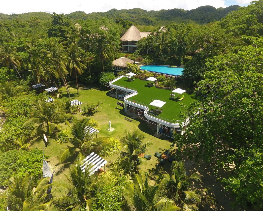 Aerial view of a lush tropical resort with green-roofed buildings, pool, and dense surrounding trees.