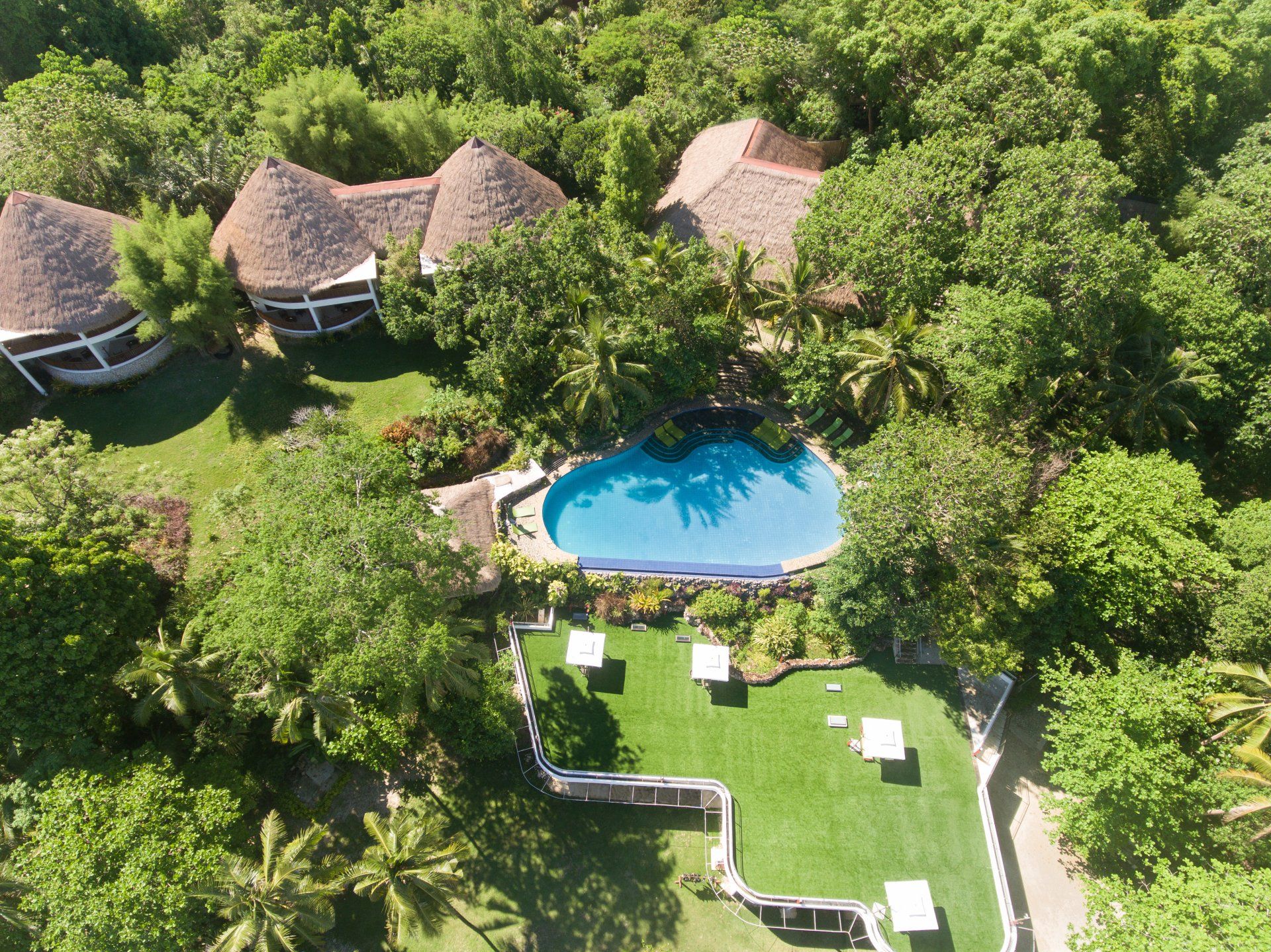 Aerial view of a tropical resort with thatched huts, a blue pool, and a green lawn surrounded by dense trees