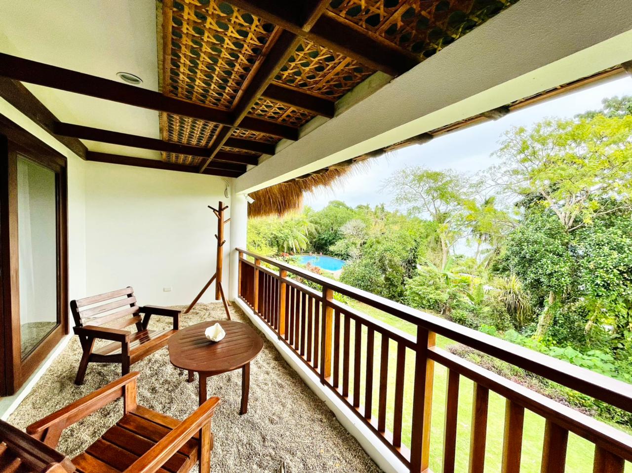 Covered balcony with wooden chairs and table overlooking a lush green landscape and pool.