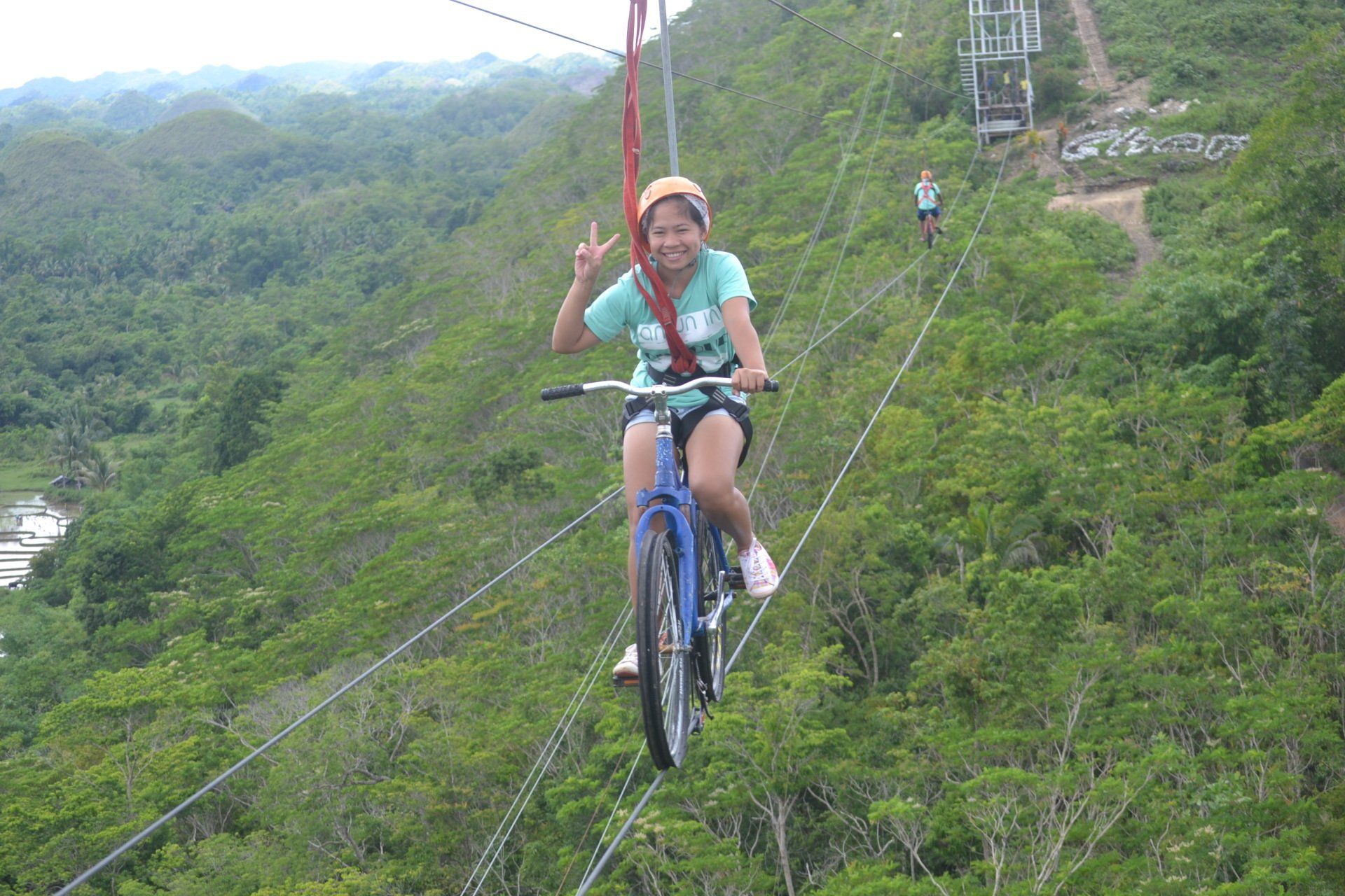 Person zip-lining over a green forest, smiling and waving on a suspended cable.