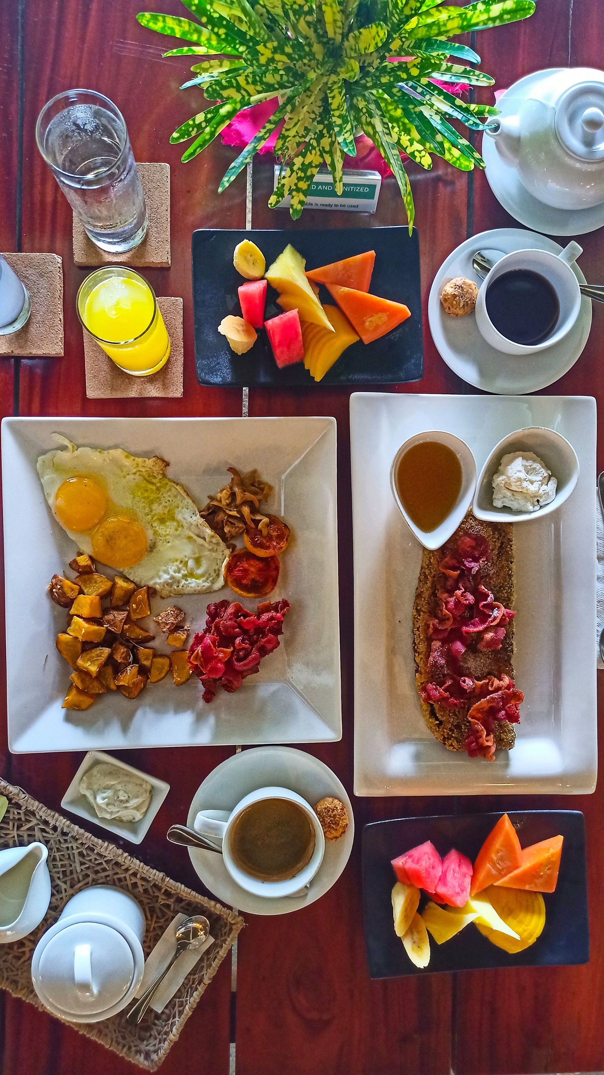 Overhead view of a colorful breakfast spread with coffee, juice, fruit, pastries, and flowers on a wooden table