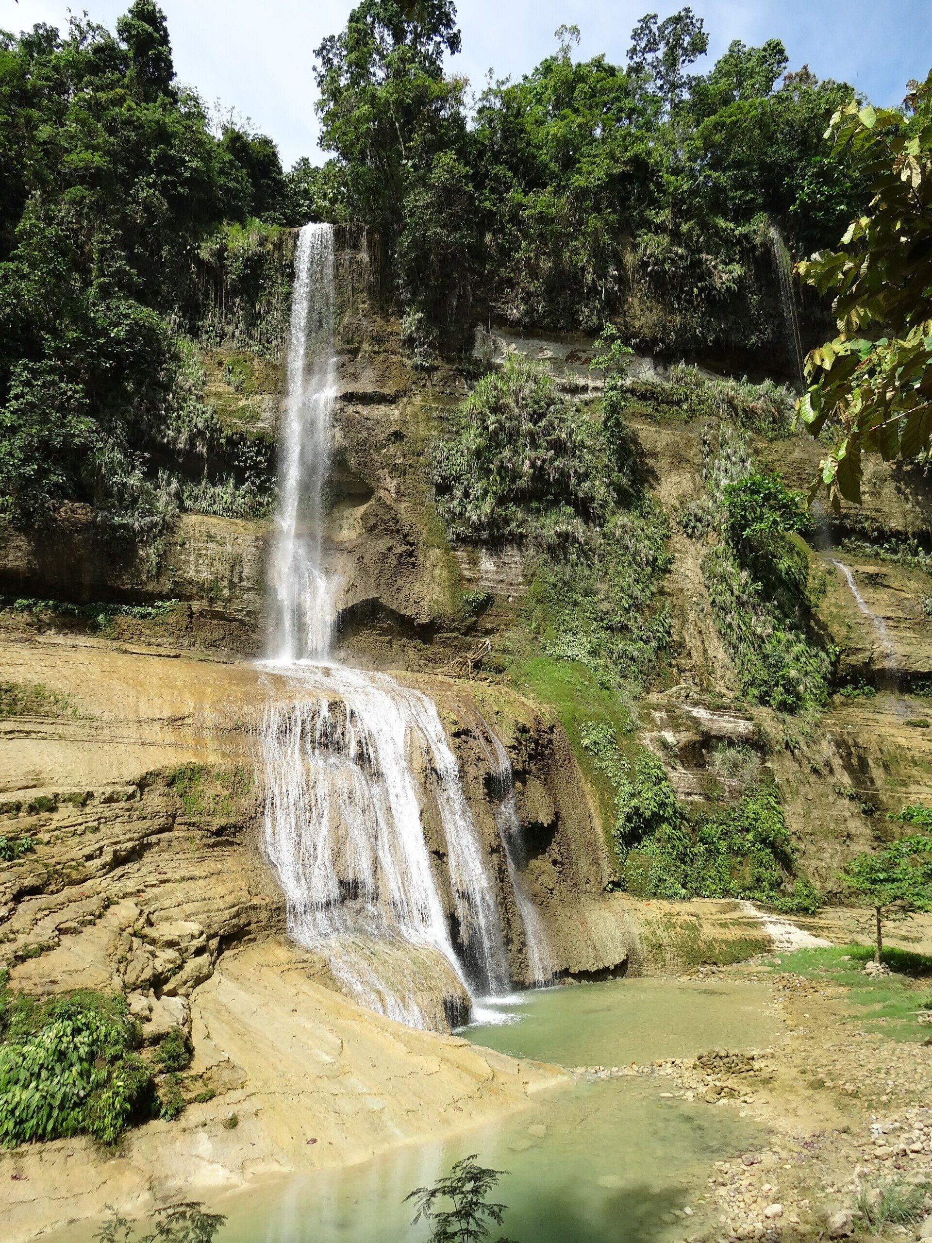 Tall waterfall cascading over a rocky cliff into a green pool, surrounded by dense forest.