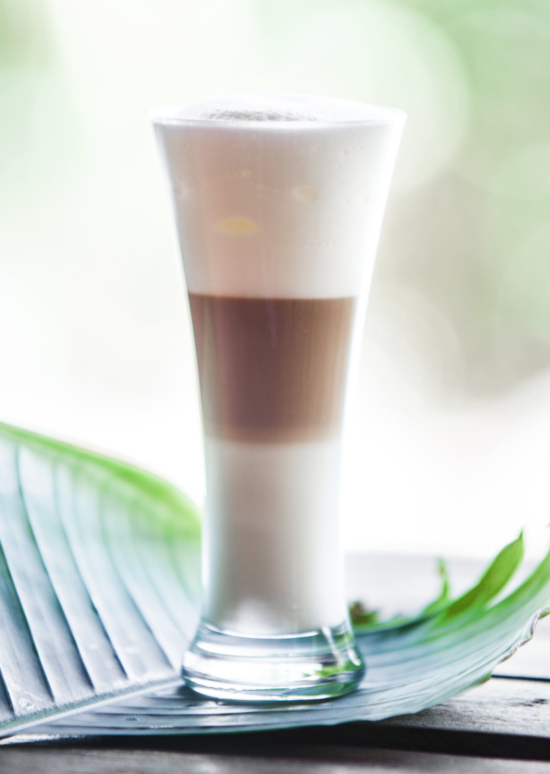 Tall layered iced drink in a clear glass on a leaf-shaped tray, with a soft bright background