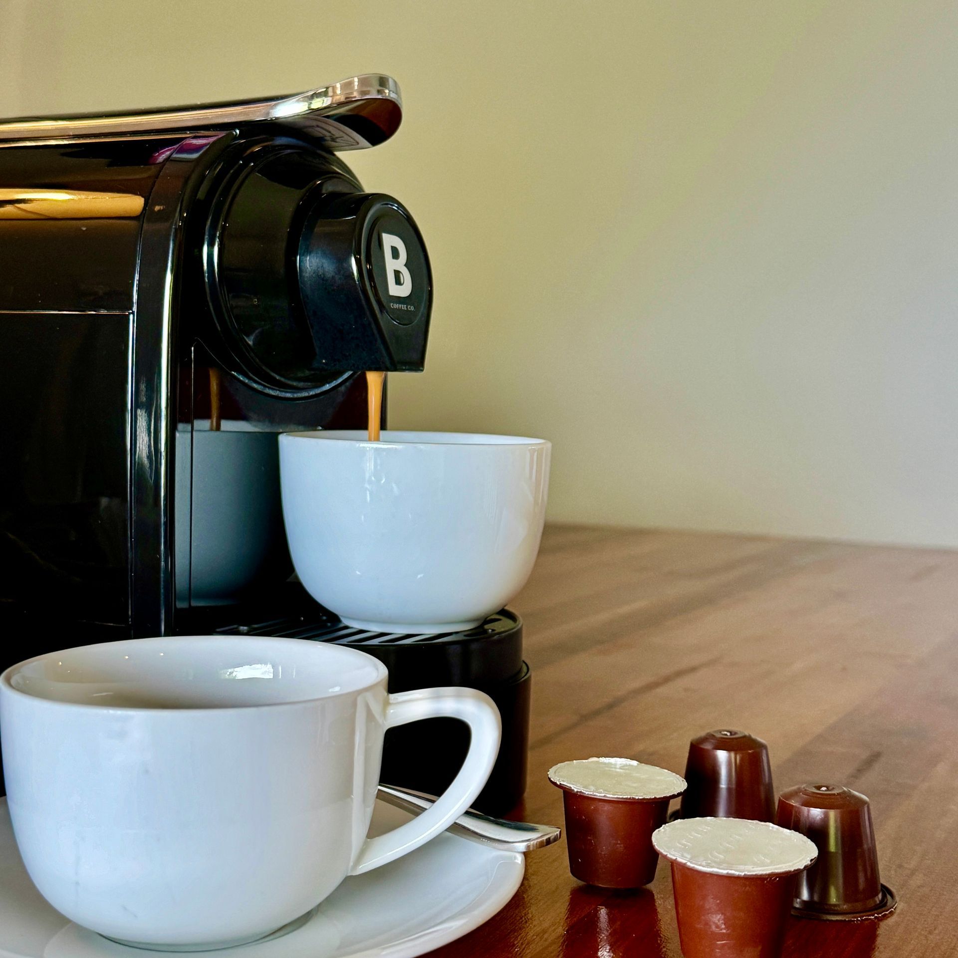 Coffee machine pouring into a white cup beside more cups and capsules on a wooden counter.