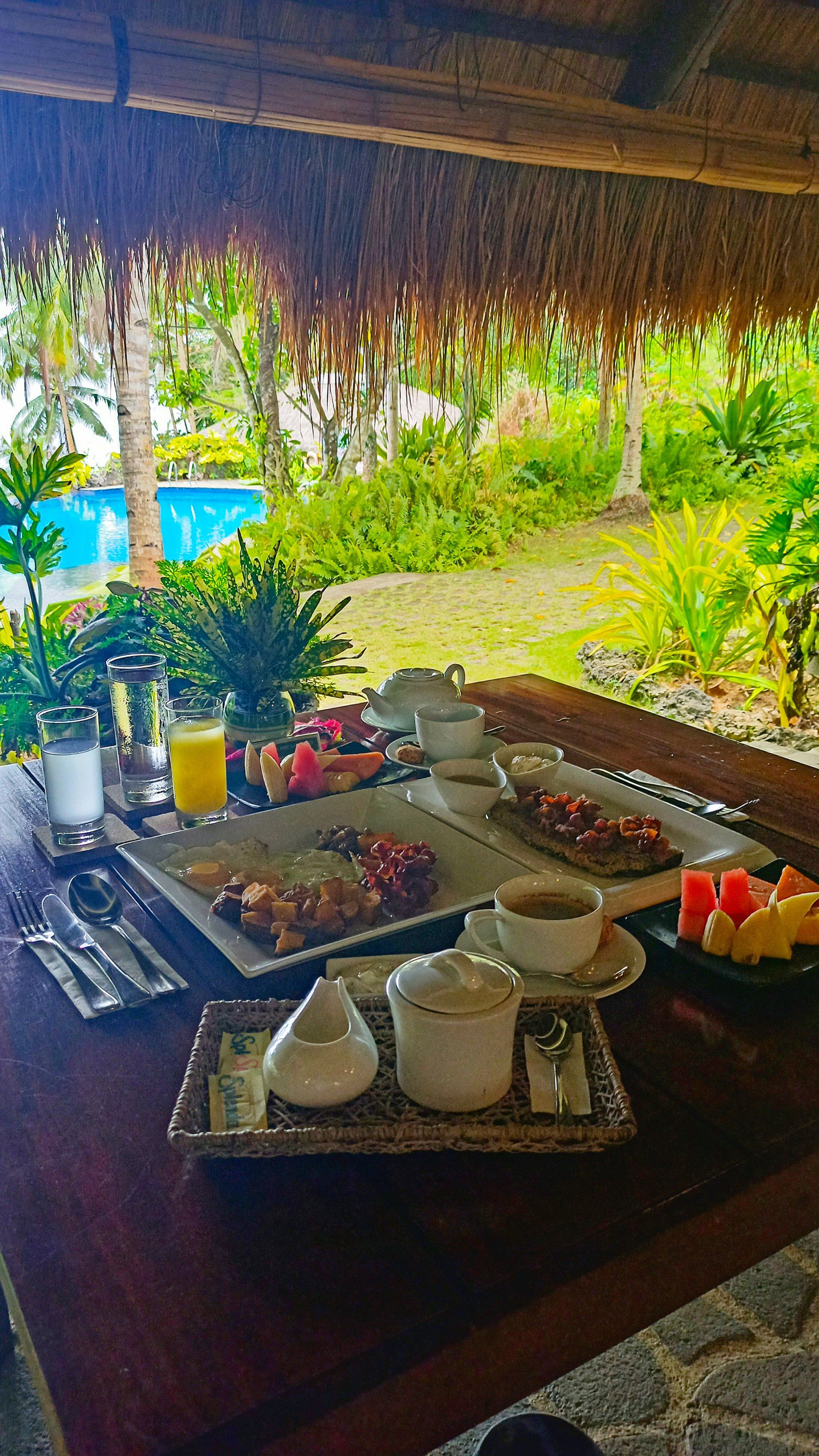 Outdoor buffet table under a thatched roof with trays of food, dishes, and condiments overlooking a lush garden