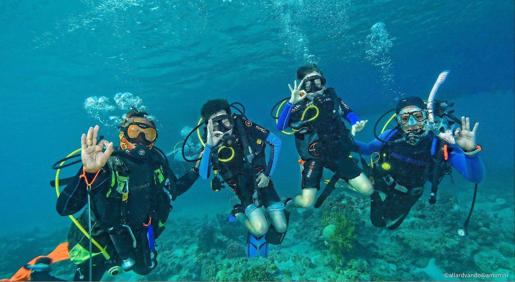 Four scuba divers underwater, posing and waving in blue open water with coral below