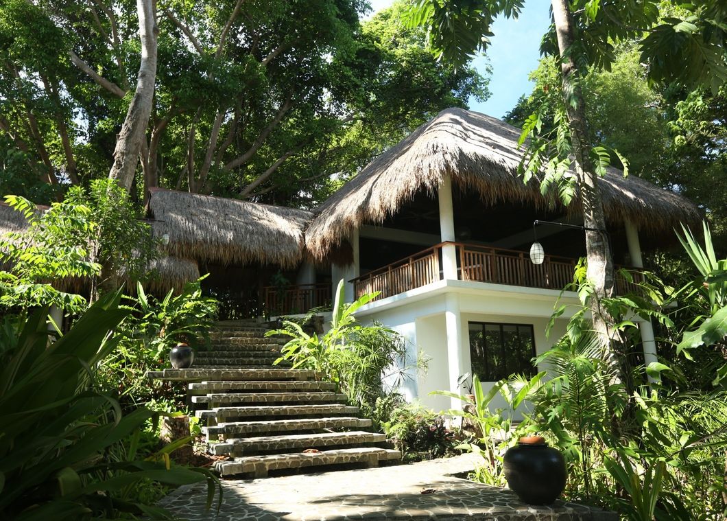 Tropical resort villa with thatched roofs, stone steps, and lush green trees surrounding it.