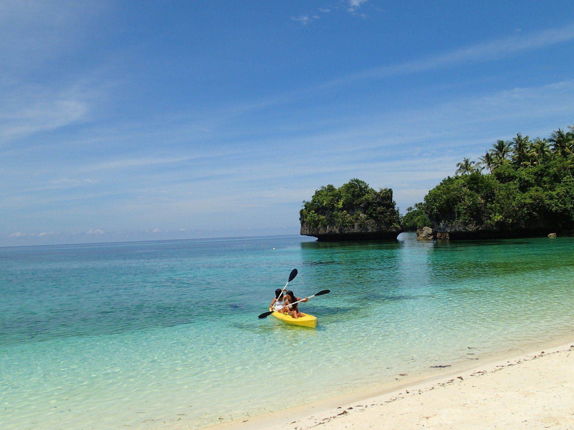 Kayaker paddles a yellow kayak in clear turquoise water near a sandy tropical beach and lush islands