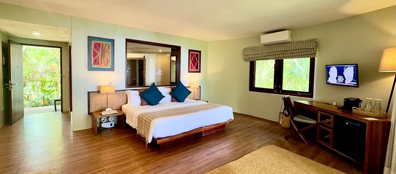 Bright tropical bedroom with king bed, wood floors, desk, TV, and open doors to greenery