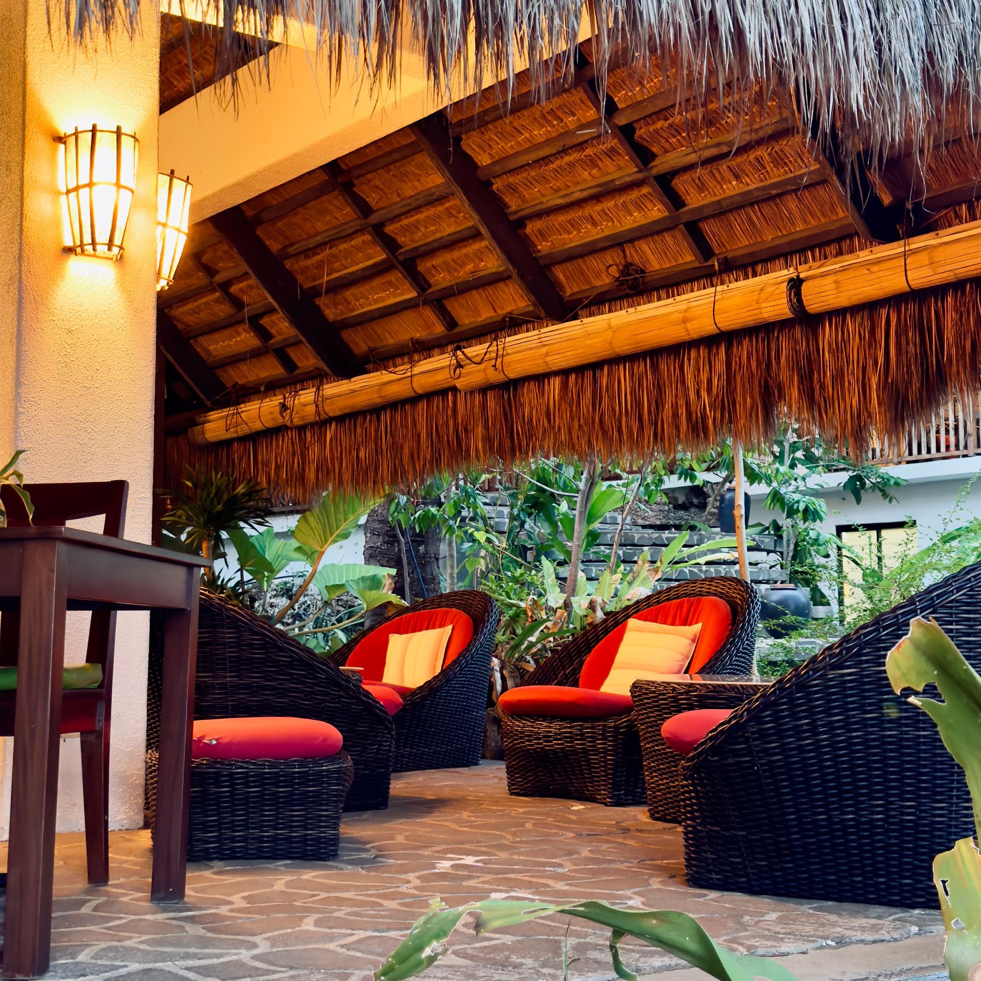 Tropical patio lounge with wicker chairs, red cushions, and a thatched roof at dusk