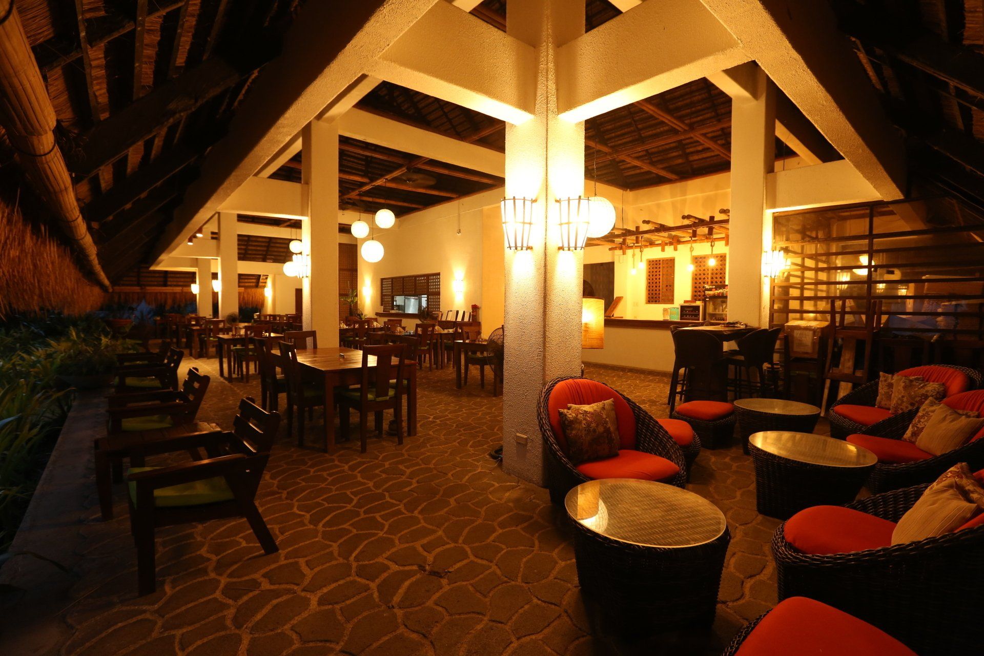 Cozy dimly lit lounge with wicker chairs, red cushions, tables, and warm yellow lighting