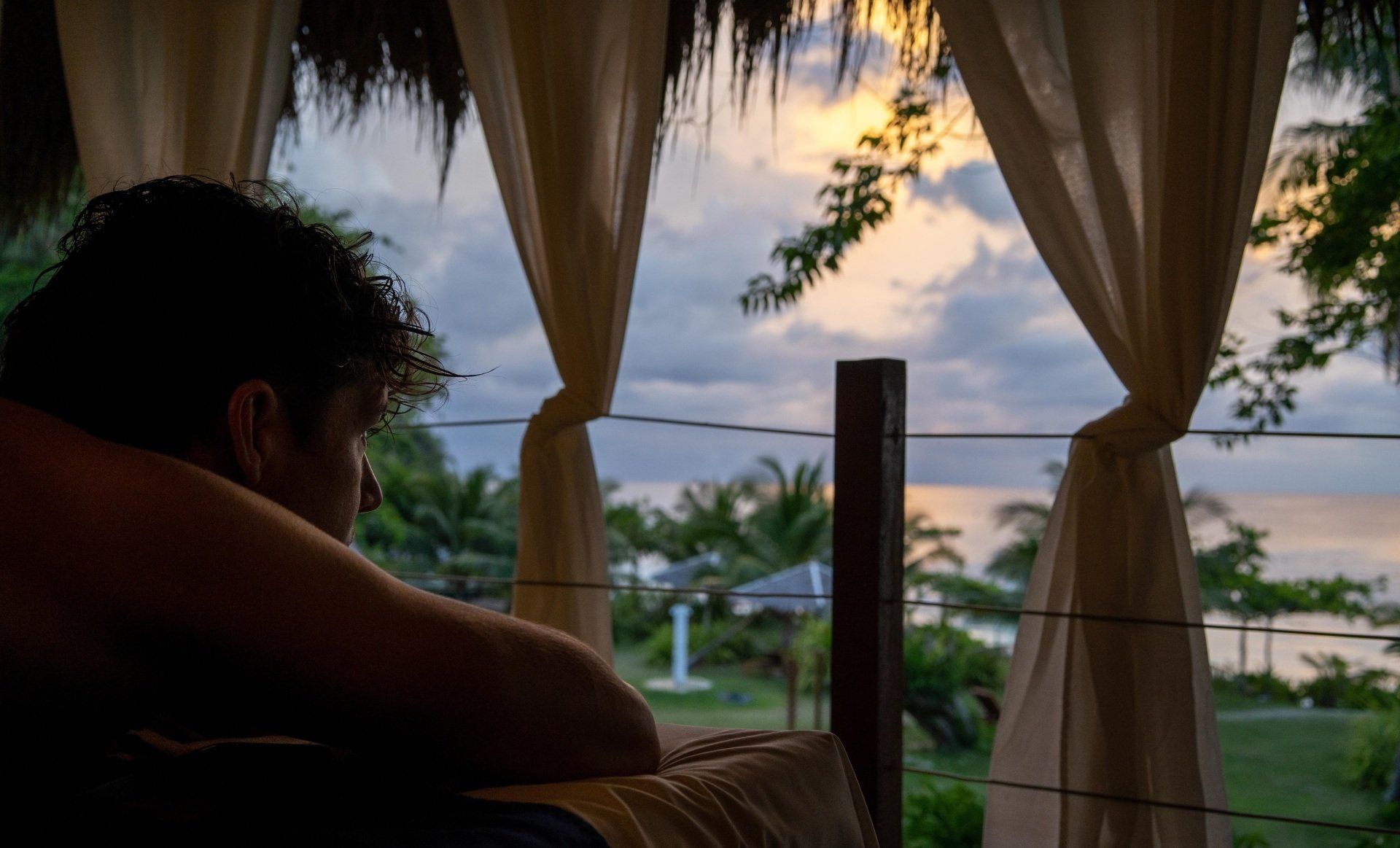 Silhouette of a person on a balcony at sunset, framed by curtains overlooking a tropical waterfront