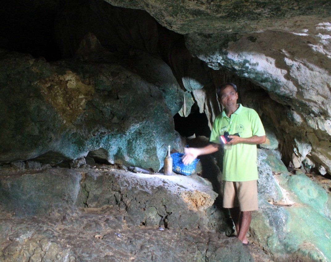 Person in a green shirt standing inside a rocky cave, holding an object near a blue container.