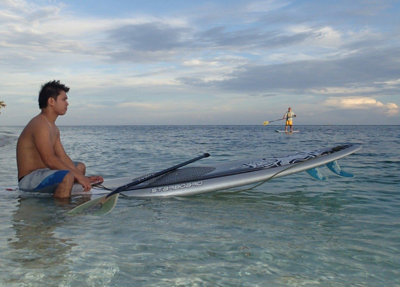 Person sitting on a surfboard in calm ocean water at sunset, with another distant paddleboarder nearby