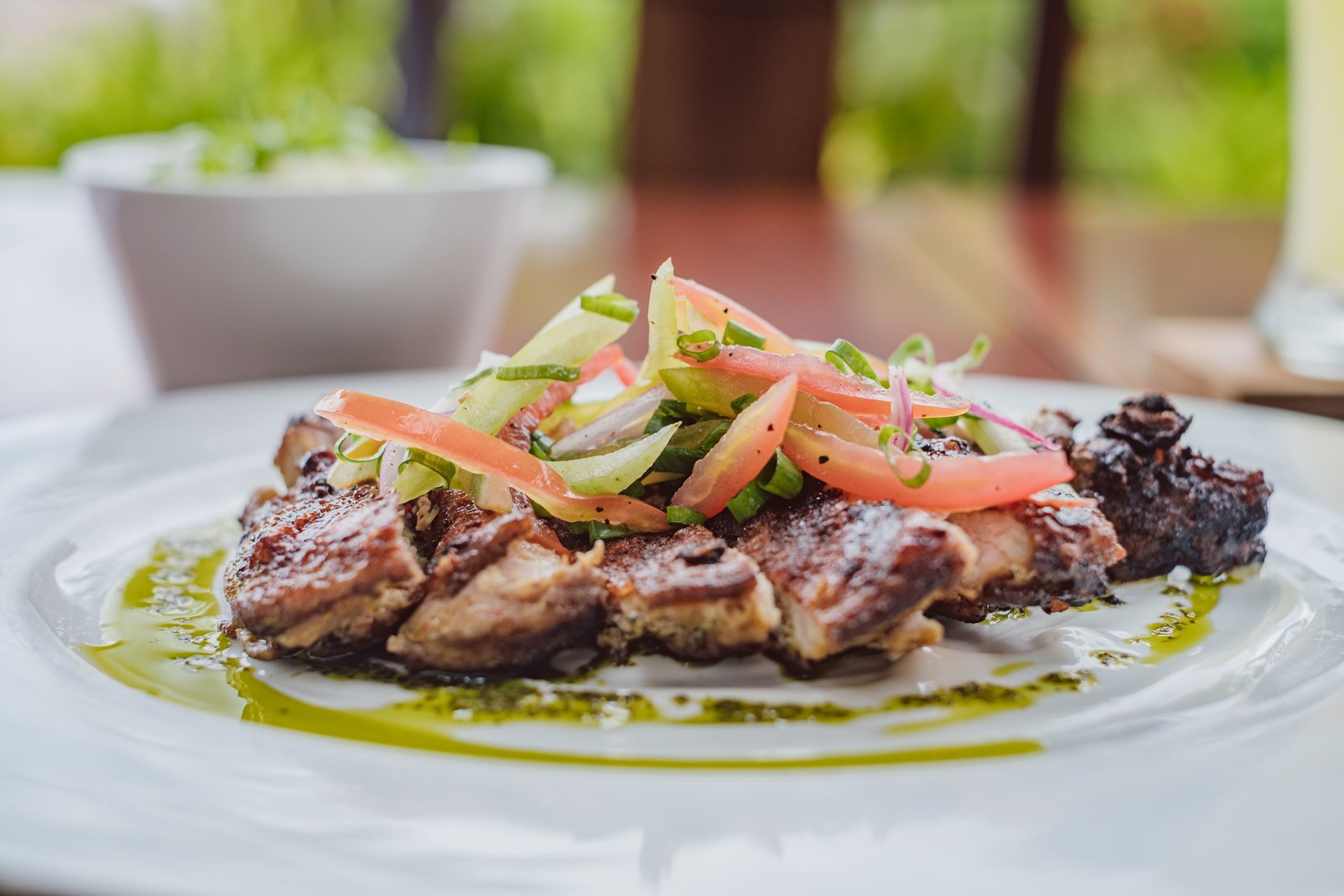 Grilled steak topped with tomato and onion salad on a white plate, drizzled with green sauce