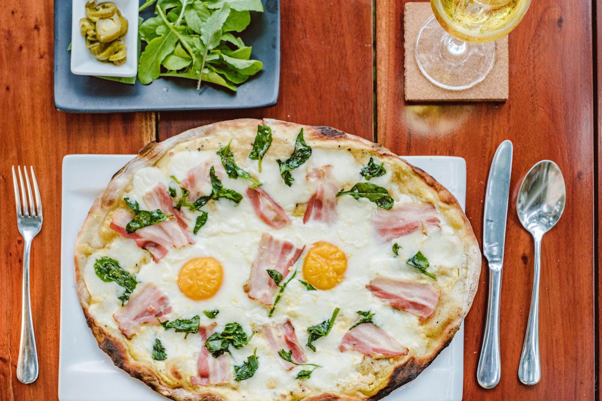 Breakfast pizza on a wooden table with two sunny-side-up eggs, bacon, greens, and side dishes
