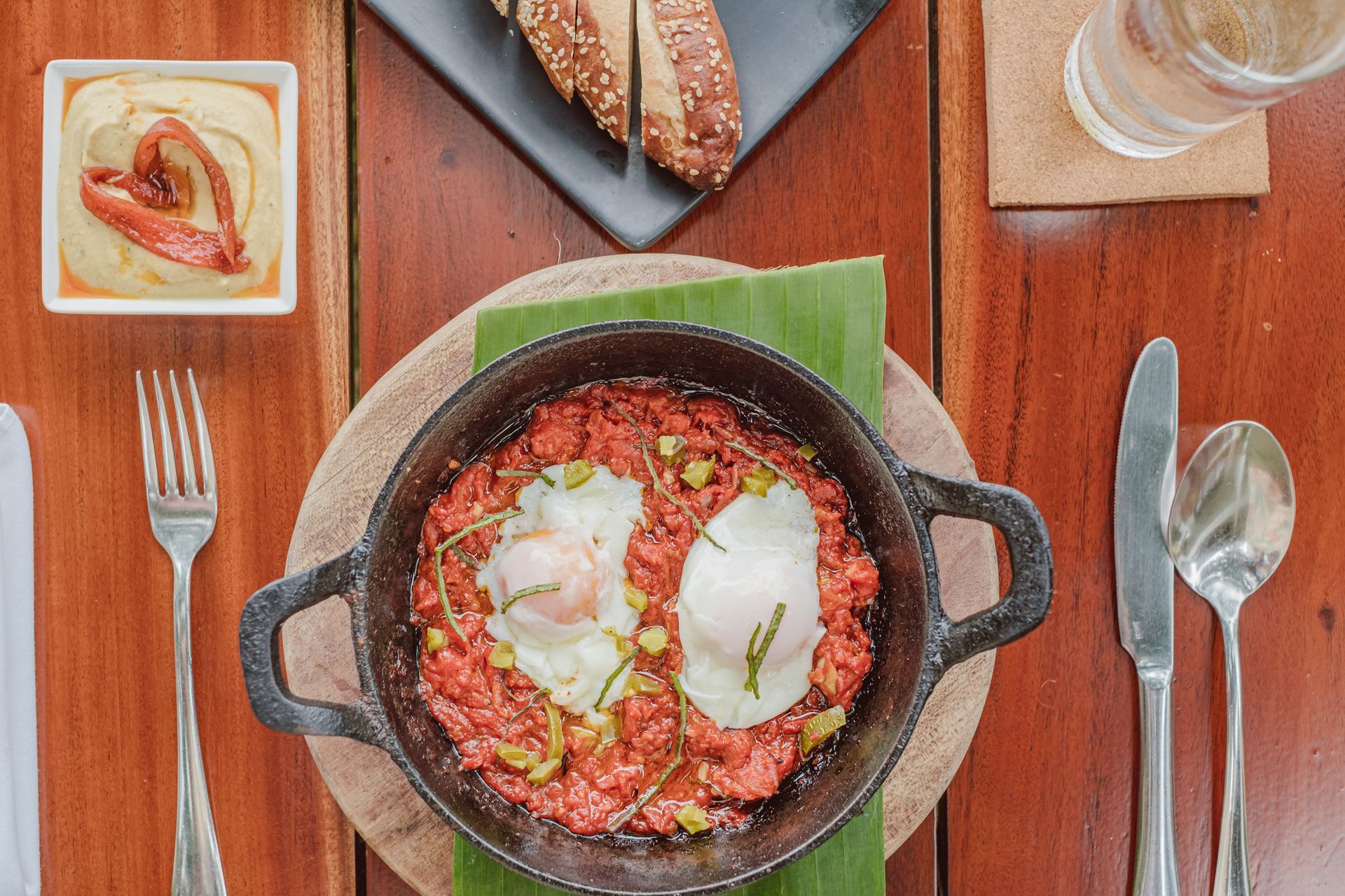 Skillet shakshuka with poached eggs on a wooden table with bread, utensils, and a glass of water